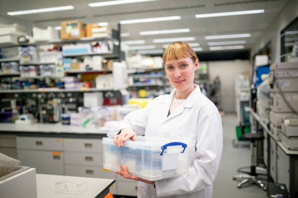 A portrait of a confident caucasian university scientist in a lab, standing tall and looking proud in her white lab coat. She is carrying a box full of documents and lab materials.