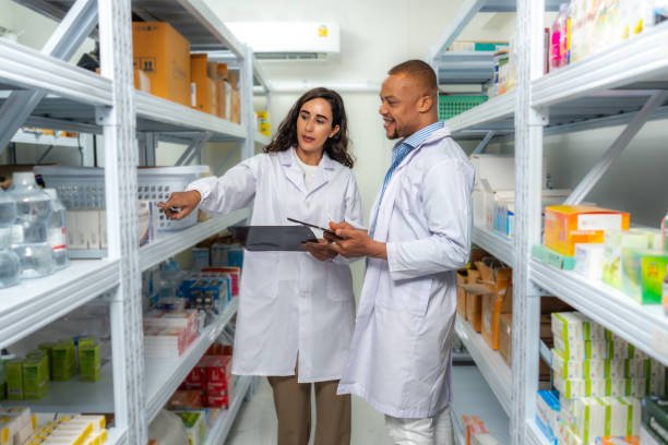 Two multiracial pharmacists, Latin female and African male working in pharmacy inventory room together by using digital tablet for checking medicine inventory storage on shelf. Storehouse supervisor female and logistic engineer male standing together at storage room.